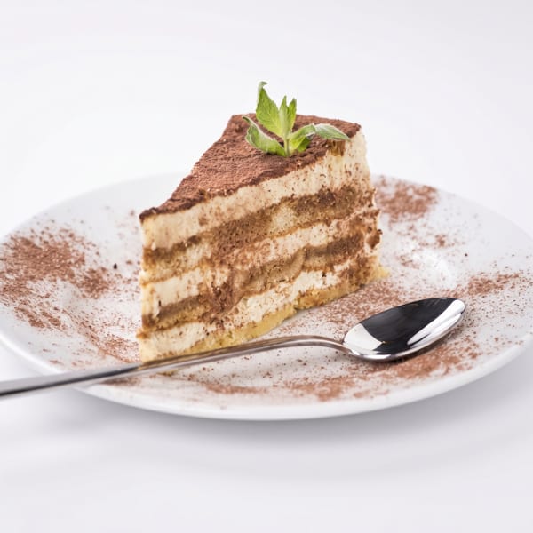 A slice of tiramisu cake with cocoa powder and a mint leaf on a white plate with a spoon.