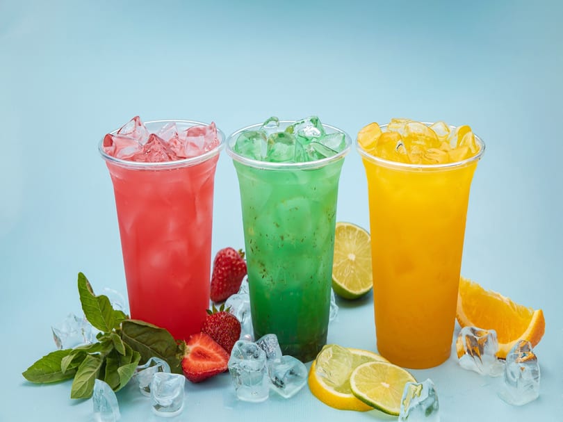 Three colorful iced drinks, red, green, and yellow, with fresh fruit and ice on a blue background.