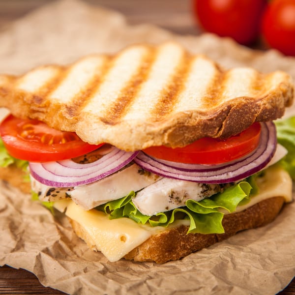 Grilled chicken sandwich with lettuce, cheese, red onion, and tomato on crumpled brown paper.