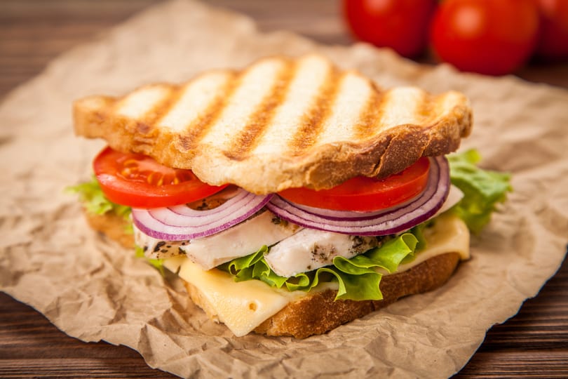 Grilled chicken sandwich with lettuce, cheese, red onion, and tomato on crumpled brown paper.
